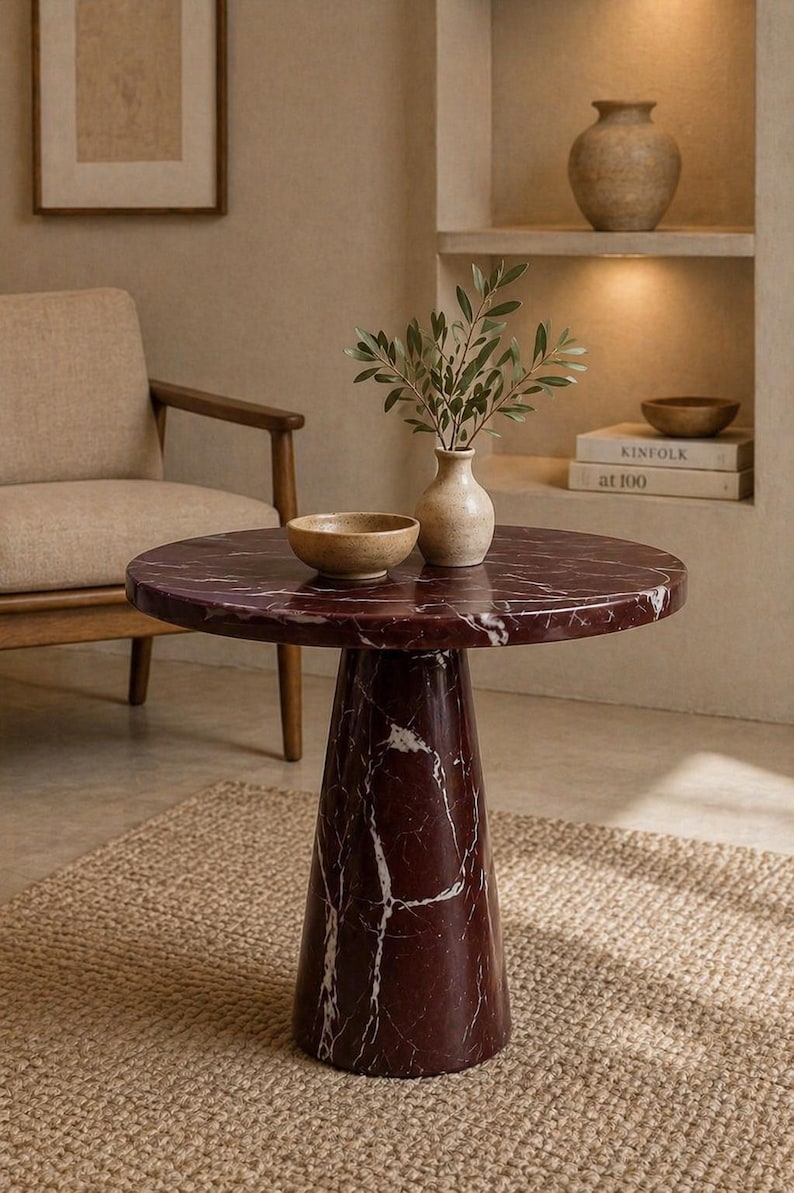 May include: A round, burgundy marble side table with white veining. The table has a single, conical base. A small vase with greenery and a wooden bowl sit on top. The table is on a woven rug.