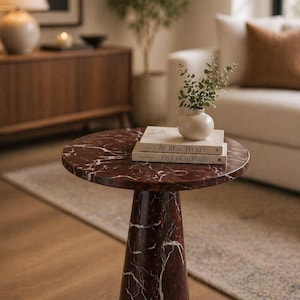 May include: A round, red marble side table with white veining. The table has a conical base and holds two books titled "Live Beautifully" and a small vase with greenery. The table is on a wooden floor.