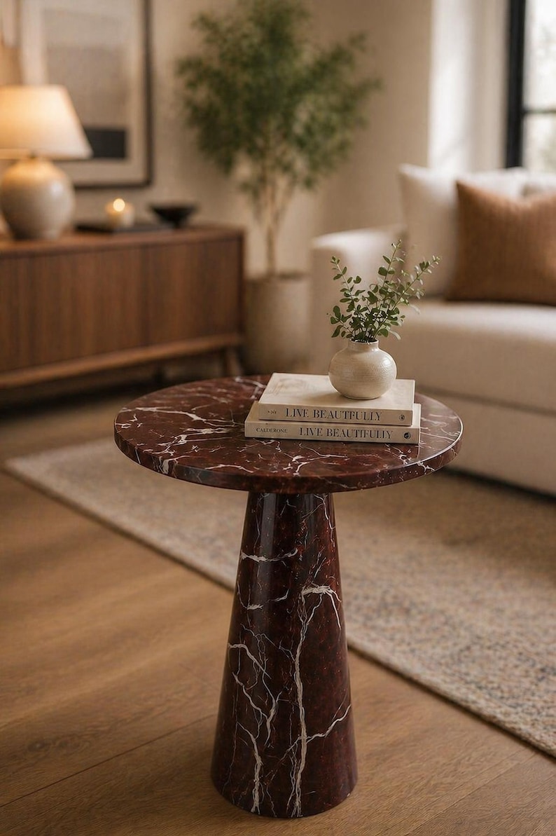 May include: A round, red marble side table with white veining. The table has a conical base and holds two books titled "Live Beautifully" and a small vase with greenery. The table is on a wooden floor.