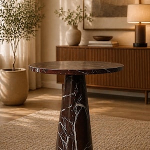 May include: A round, dark brown marble side table with white veining. The table has a conical base and sits on a neutral-colored rug. The table is approximately 24 inches in diameter.