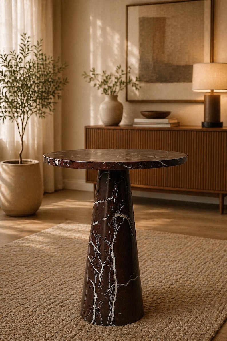 May include: A round, dark brown marble side table with white veining. The table has a conical base and sits on a neutral-colored rug. The table is approximately 24 inches in diameter.
