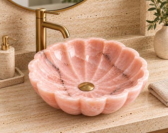 May include: A round, scalloped-edge bathroom sink carved from polished pink stone with natural dark veining. The basin features a central gold-toned drain cover and is paired with a matching gold-toned faucet fixture.