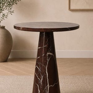 May include: A round, dark brown marble side table with white veining. The table has a conical base and a circular top. The table is set on a neutral-colored rug and is a decorative accent piece.