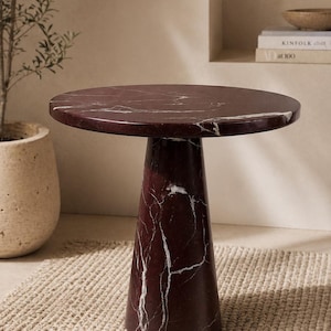May include: A round, burgundy marble side table with white veining. The table has a cylindrical base that flares out slightly towards the bottom. The table is set on a woven rug, with a potted plant and books in the background.