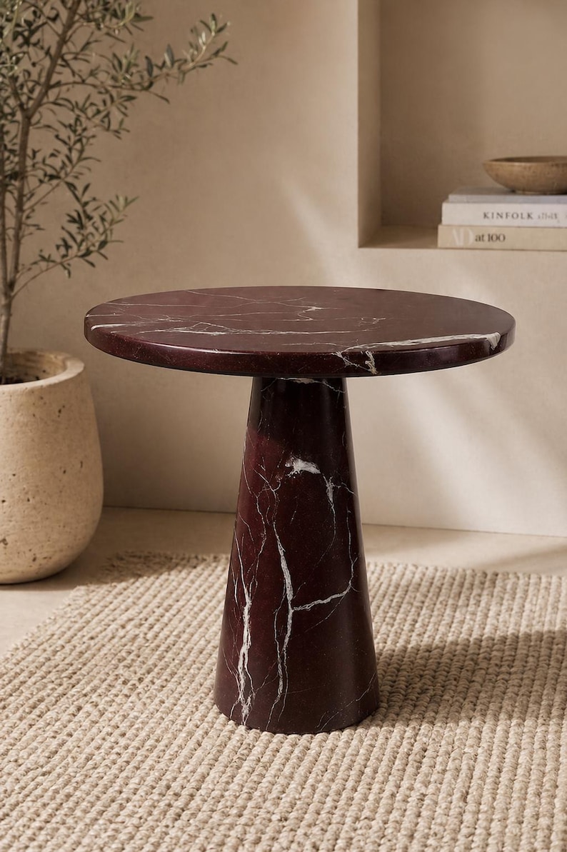 May include: A round, burgundy marble side table with white veining. The table has a cylindrical base that flares out slightly towards the bottom. The table is set on a woven rug, with a potted plant and books in the background.