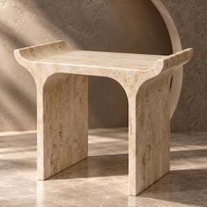 Puede incluir: Un taburete de travertino beige con una textura de piedra natural porosa. La pieza presenta un asiento rectangular con extremos ligeramente curvados hacia arriba y dos soportes laterales sólidos y arqueados que crean una silueta continua.