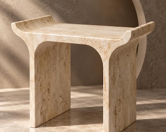 May include: A beige travertine stool with a porous, natural stone texture. The piece features a rectangular seat with slightly upturned ends and two solid, arched side supports that create a continuous, curved silhouette from top to base.