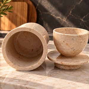 Puede incluir: Un recipiente cilíndrico de mármol beige con un patrón moteado, colocado junto a un cuenco de mármol a juego que descansa sobre una base circular de mármol. Ambos artículos presentan un acabado de piedra natural pulida con vetas visibles.
