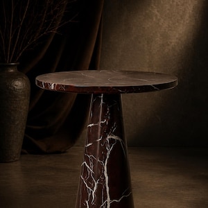 May include: A round, dark red marble side table with white veining. The table has a conical base and a circular top. The table is approximately 20 inches in diameter and 24 inches tall. The background is a dark brown.