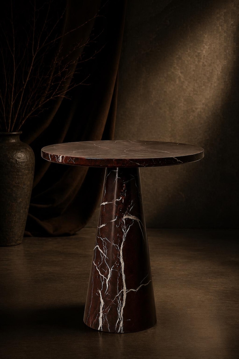 May include: A round, dark red marble side table with white veining. The table has a conical base and a circular top. The table is approximately 20 inches in diameter and 24 inches tall. The background is a dark brown.