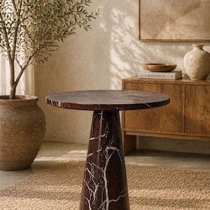 May include: A round, dark brown marble side table with white veining. The table has a conical base and sits on a woven jute rug. A potted olive tree and wooden cabinet are in the background.