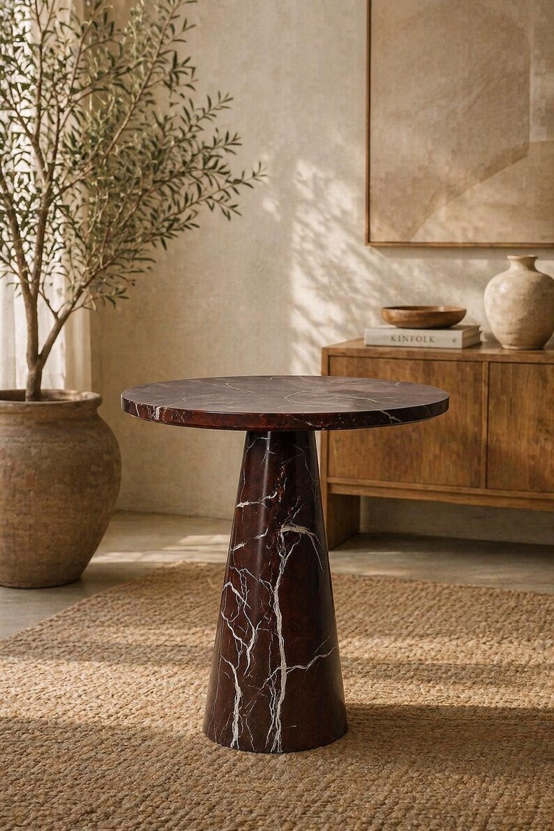 May include: A round, dark brown marble side table with white veining. The table has a conical base and sits on a woven jute rug. A potted olive tree and wooden cabinet are in the background.