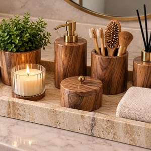 May include: A rectangular beige stone tray holds a collection of bathroom accessories including a wood-grain soap dispenser, a lidded jar, a toothbrush holder with brushes, a small potted plant, a lit candle in a glass, and a folded towel.