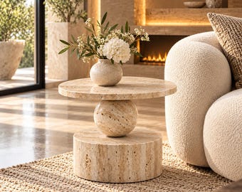 May include: A round side table crafted from beige travertine stone, featuring a flat circular top supported by a central spherical element resting on a thick cylindrical base. A small matching travertine vase holds white flowers.
