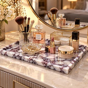 Puede incluir: Una bandeja rectangular de mármol blanco y morado que contiene un portaescobillas de cristal con brochas de maquillaje, un frasco de perfume Coco Mademoiselle, un pequeño plato de cristal con joyas, una caja de cristal dorada y labial.