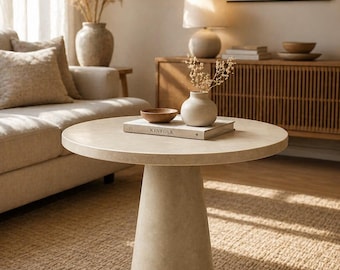 May include: A neutral-toned living room scene featuring a round, beige coffee table with a conical base. A book titled "KINFOLK" rests on the table, alongside a small vase and a wooden bowl. The room includes a beige sofa, a rug, and decorative vases.
