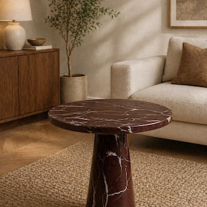May include: A round, burgundy marble side table with white veining. The table has a conical base and sits on a woven jute rug. The background includes a sofa, a wooden cabinet, and a potted plant, creating a warm, inviting living room setting.