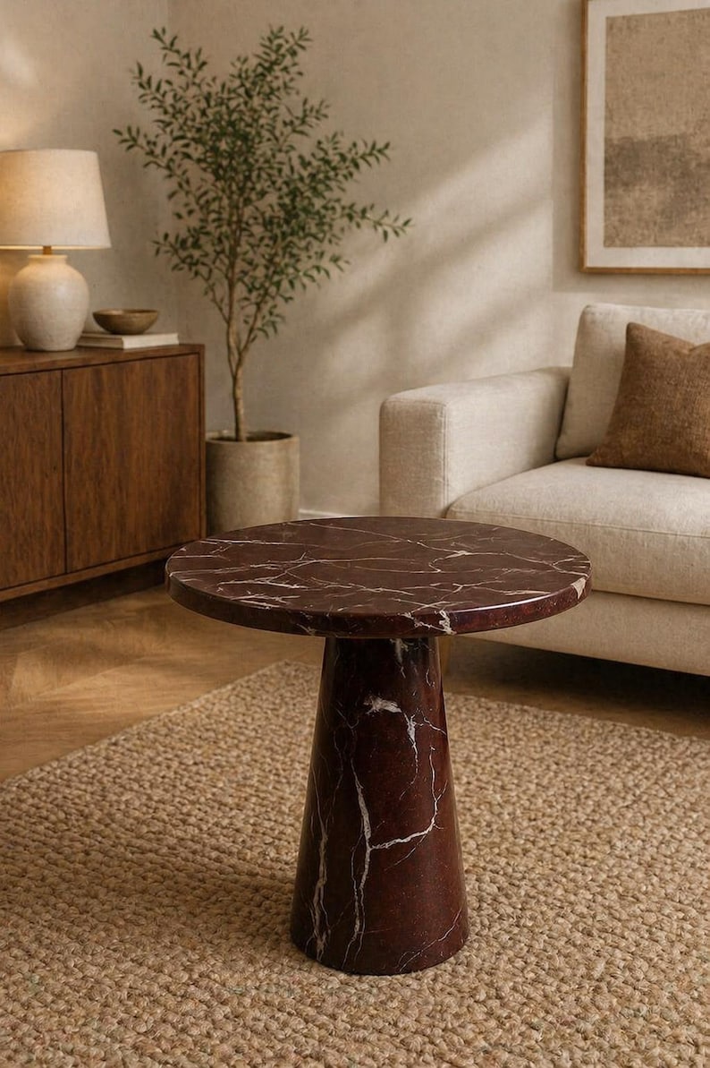May include: A round, burgundy marble side table with white veining. The table has a conical base and sits on a woven jute rug. The background includes a sofa, a wooden cabinet, and a potted plant, creating a warm, inviting living room setting.