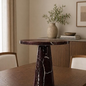 May include: A round, dark red marble side table with white veining. The table has a conical base and sits on a dark wood table. The room has a neutral color palette with a vase of greenery and a framed picture.