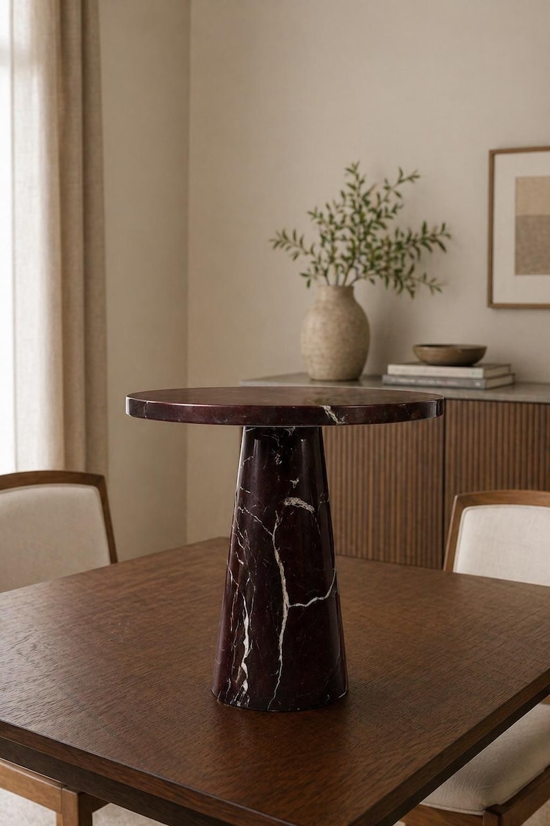May include: A round, dark red marble side table with white veining. The table has a conical base and sits on a dark wood table. The room has a neutral color palette with a vase of greenery and a framed picture.