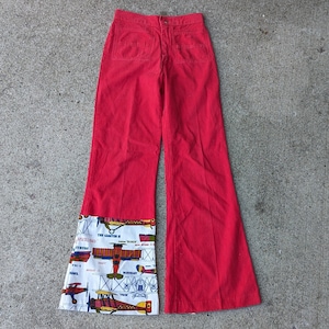 May include: Bright red flared pants with a white and patterned fabric detail at the hem. The patterned fabric features vintage airplane illustrations and text. The pants have two back pockets and a button closure.