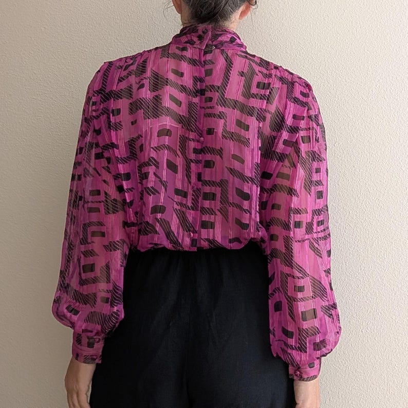 May include: A sheer, long-sleeved blouse in a vibrant magenta hue, patterned with black geometric shapes. The blouse features a high neckline and gathered cuffs, paired with black bottoms. The fabric appears lightweight and slightly textured.