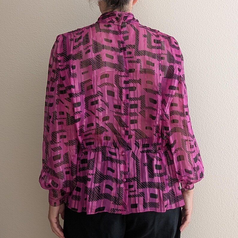 May include: A sheer magenta blouse with a black geometric pattern. The long-sleeved top has a high collar, gathered waist, and button details. The blouse is made of a lightweight, semi-transparent fabric.