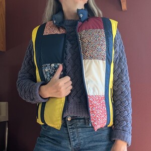May include: A yellow quilted vest with a patchwork design. The vest has a zipper closure and is made of different fabrics with floral patterns. The vest is worn over a gray sweater.