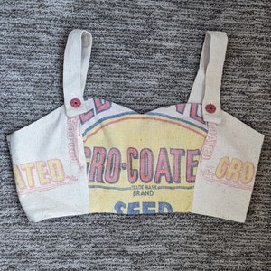 May include: A white fabric crop top with adjustable straps. The fabric has a vintage look and features the text "Gro-Coated Seed" in red and yellow. The straps have red buttons.