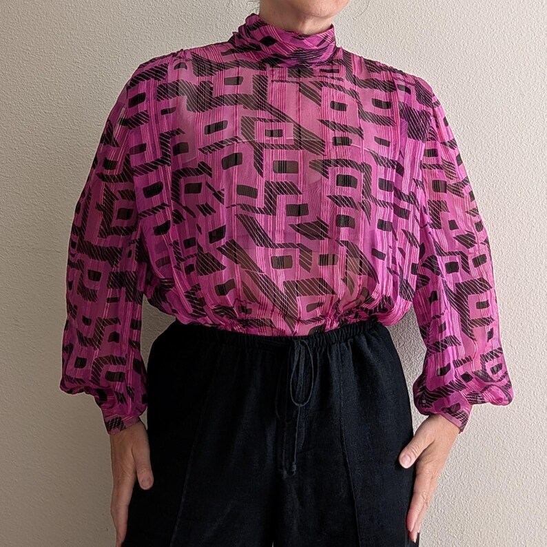 May include: A pink blouse with a high neck and long sleeves. The blouse features a geometric black and pink pattern. The bottom of the blouse is gathered at the waist. The person in the image is wearing black pants.