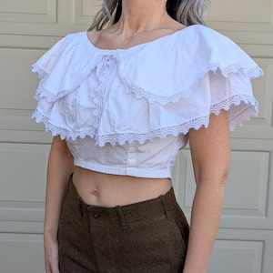 May include: White crop top with a large, ruffled collar and lace trim. The top is made of a lightweight fabric and has a fitted bodice.