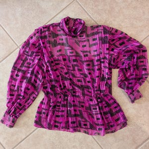 May include: A magenta blouse with a black geometric pattern. The blouse features a high neckline, long, voluminous sleeves, and a gathered waist. The fabric appears sheer and lightweight.