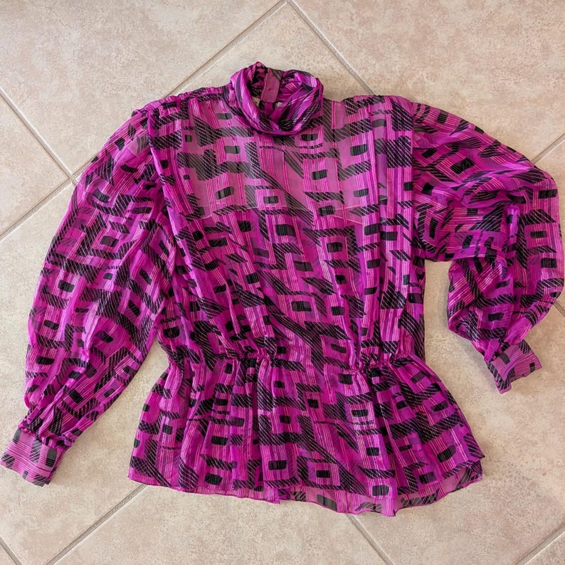 May include: A magenta blouse with a black geometric pattern. The blouse features a high neckline, long, voluminous sleeves, and a gathered waist. The fabric appears sheer and lightweight.