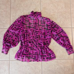 May include: A bright pink blouse with a high collar and long sleeves. The blouse features a black geometric pattern and a cinched waist. The fabric appears to be sheer and lightweight, with a slight sheen.
