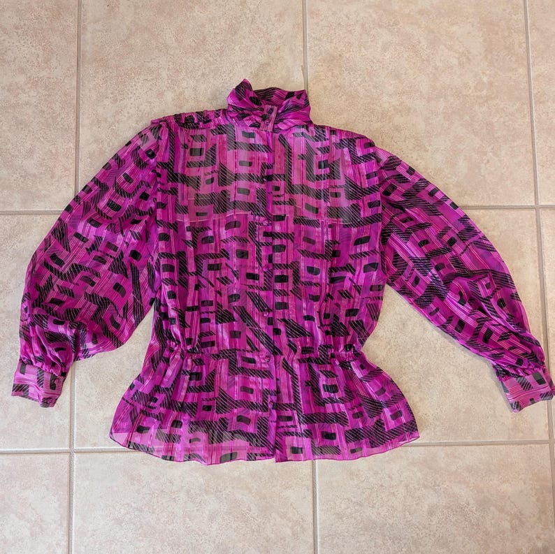 May include: A bright pink blouse with a high collar and long sleeves. The blouse features a black geometric pattern and a cinched waist. The fabric appears to be sheer and lightweight, with a slight sheen.