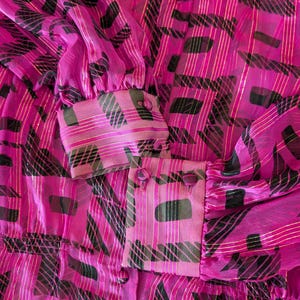 May include: Close-up of a vibrant pink blouse with a semi-sheer fabric. The blouse features a repeating pattern of black and green shapes, accented with gold stripes. The cuffs have pink buttons and a matching pattern.