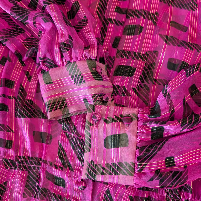 May include: Close-up of a vibrant pink blouse with a semi-sheer fabric. The blouse features a repeating pattern of black and green shapes, accented with gold stripes. The cuffs have pink buttons and a matching pattern.