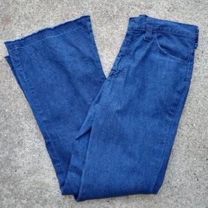 May include: Blue denim bell-bottom jeans with a frayed hem. The jeans have a classic five-pocket design and a button and zipper closure. The fabric appears to be a medium-weight denim.
