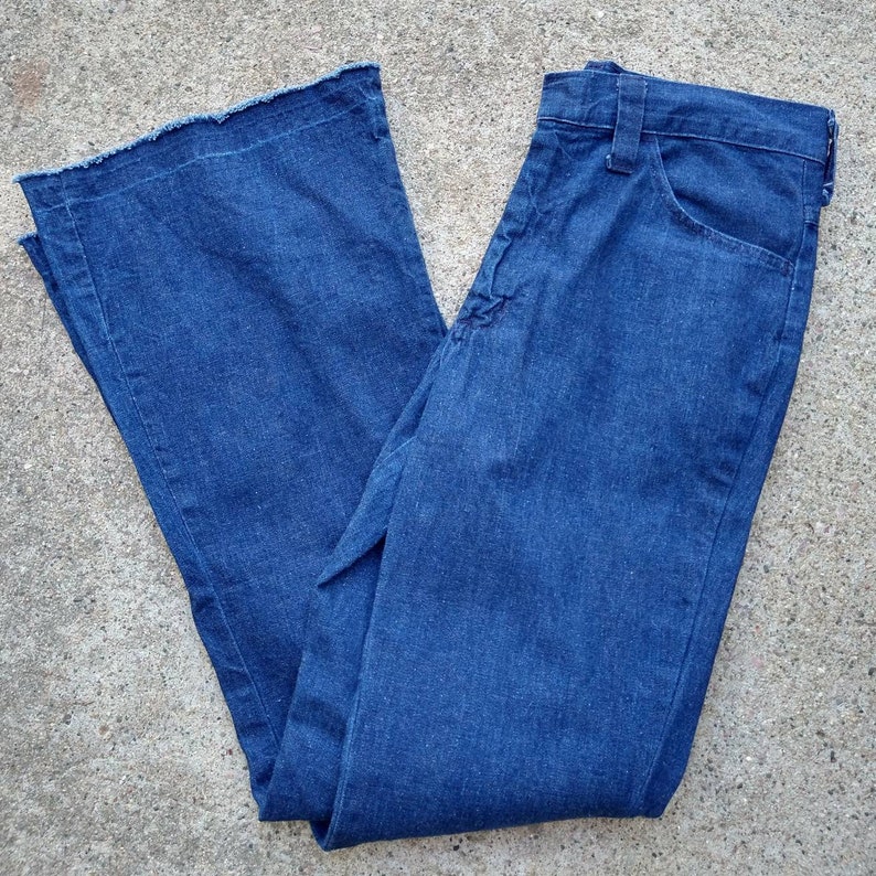 May include: Blue denim bell-bottom jeans with a frayed hem. The jeans have a classic five-pocket design and a button and zipper closure. The fabric appears to be a medium-weight denim.