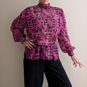 May include: A sheer, magenta blouse with a high neckline and long sleeves. The blouse features a geometric black pattern and a gathered waist. The sleeves have a slight puff at the shoulder and gather at the wrist. The blouse is paired with black pants.