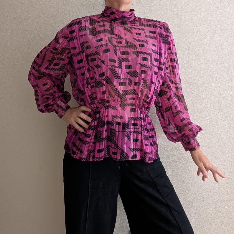 May include: A sheer, magenta blouse with a high neckline and long sleeves. The blouse features a geometric black pattern and a gathered waist. The sleeves have a slight puff at the shoulder and gather at the wrist. The blouse is paired with black pants.