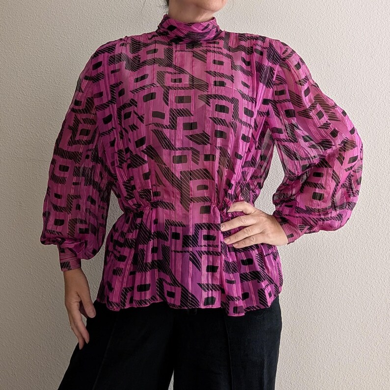 May include: A sheer, magenta blouse with a high neckline and long, puffed sleeves. The blouse features a geometric black pattern and a cinched waist. The garment is paired with black pants.
