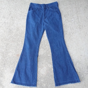 May include: Blue denim bell-bottom pants. The pants have a classic five-pocket design with a button and zipper closure. The flared legs have a raw hem. The pants are laid flat on a concrete surface.