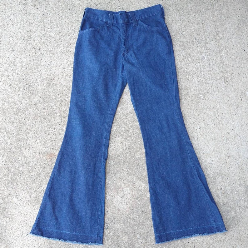 May include: Blue denim bell-bottom pants. The pants have a classic five-pocket design with a button and zipper closure. The flared legs have a raw hem. The pants are laid flat on a concrete surface.
