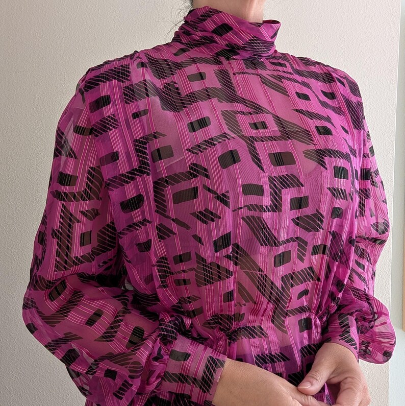 May include: A sheer, long-sleeved magenta blouse with a high neckline. The blouse features a repeating geometric pattern in black. The fabric appears lightweight and slightly textured, with a gathered waist and sleeves.