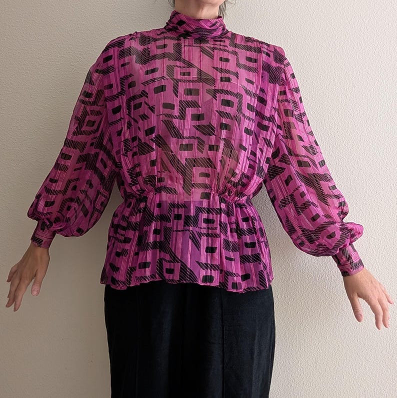 May include: A sheer, magenta blouse with a high neckline and long, voluminous sleeves. The blouse features a black geometric pattern and is gathered at the waist. The sleeves have fitted cuffs. The blouse is paired with a black skirt.