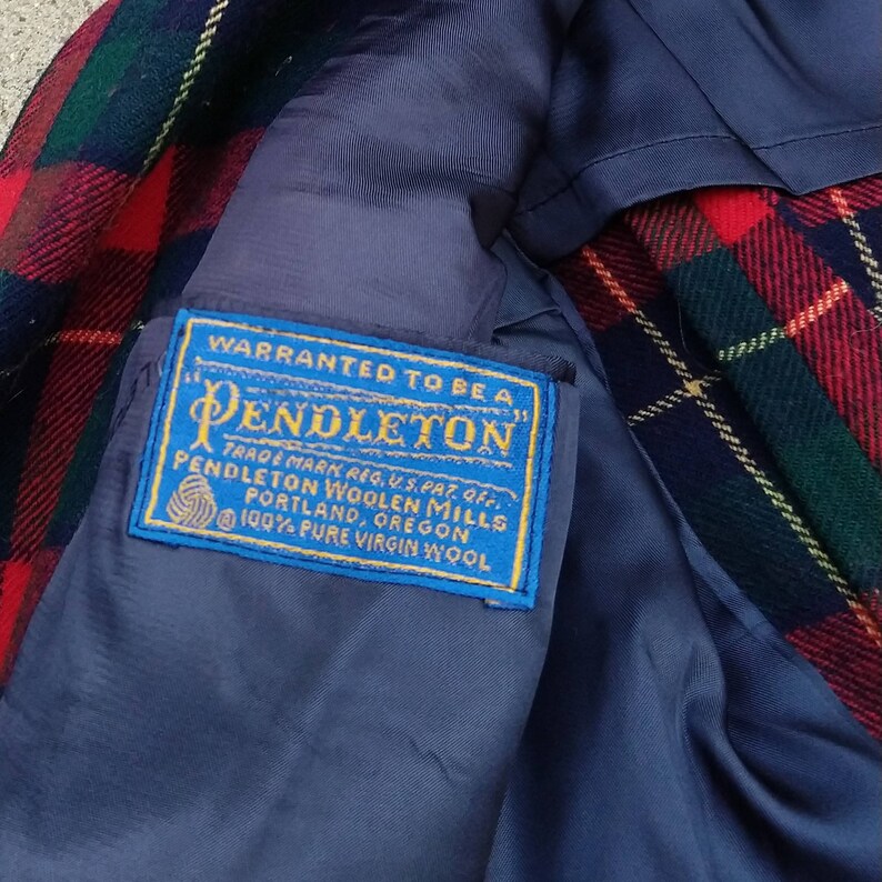 May include: A blue label with white text that reads "WARRANTED TO BE A PENDLETON" TRADE MARK REG. U.S. PAT. OFF. PENDLETON WOOLEN MILLS PORTLAND, OREGON 100% PURE VIRGIN WOOL. The label is attached to a red, green, and blue plaid fabric.