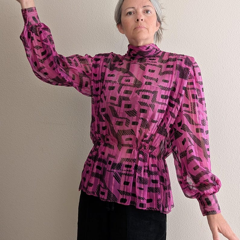 May include: A sheer, magenta blouse with a high neckline and long, voluminous sleeves. The blouse features a black geometric pattern and a cinched waist. The fabric has a pleated texture, adding dimension to the garment. The blouse is paired with a black skirt.
