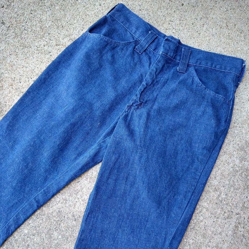 May include: A pair of blue denim jeans. The jeans have a classic five-pocket design with a button and zipper closure. The denim is a medium wash, and the jeans appear to be vintage.