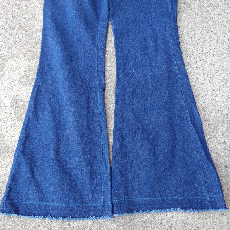 May include: A pair of blue denim bell-bottom pants. The pants have a classic five-pocket design and a wide, flared leg. The hemline is frayed. The pants are made of a medium-weight denim fabric.